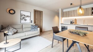 A modern living room with a sofa, coffee table, and dining table. There is a kitchen area with wooden cabinets and a hanging light fixture. - Amazing Apartment in Tyrol With Infrared Sauna (Westendorf)
