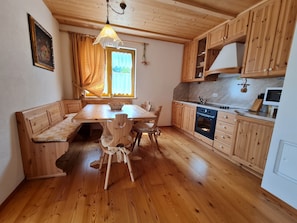 Un interior de madera con una mesa de comedor, sillas, un banco, una cocina con armarios de madera y una ventana con cortinas.