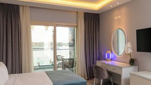 Habitación de hotel con cama, escritorio, silla, espejo, televisor y vista hacia una masa de agua a través de la ventana.