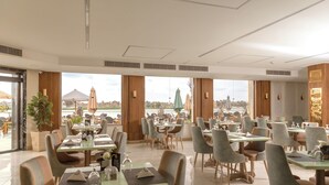 Un restaurante espacioso con varias mesas y sillas, ventanas amplias que dan a una masa de agua y edificios, y una decoración interior moderna.