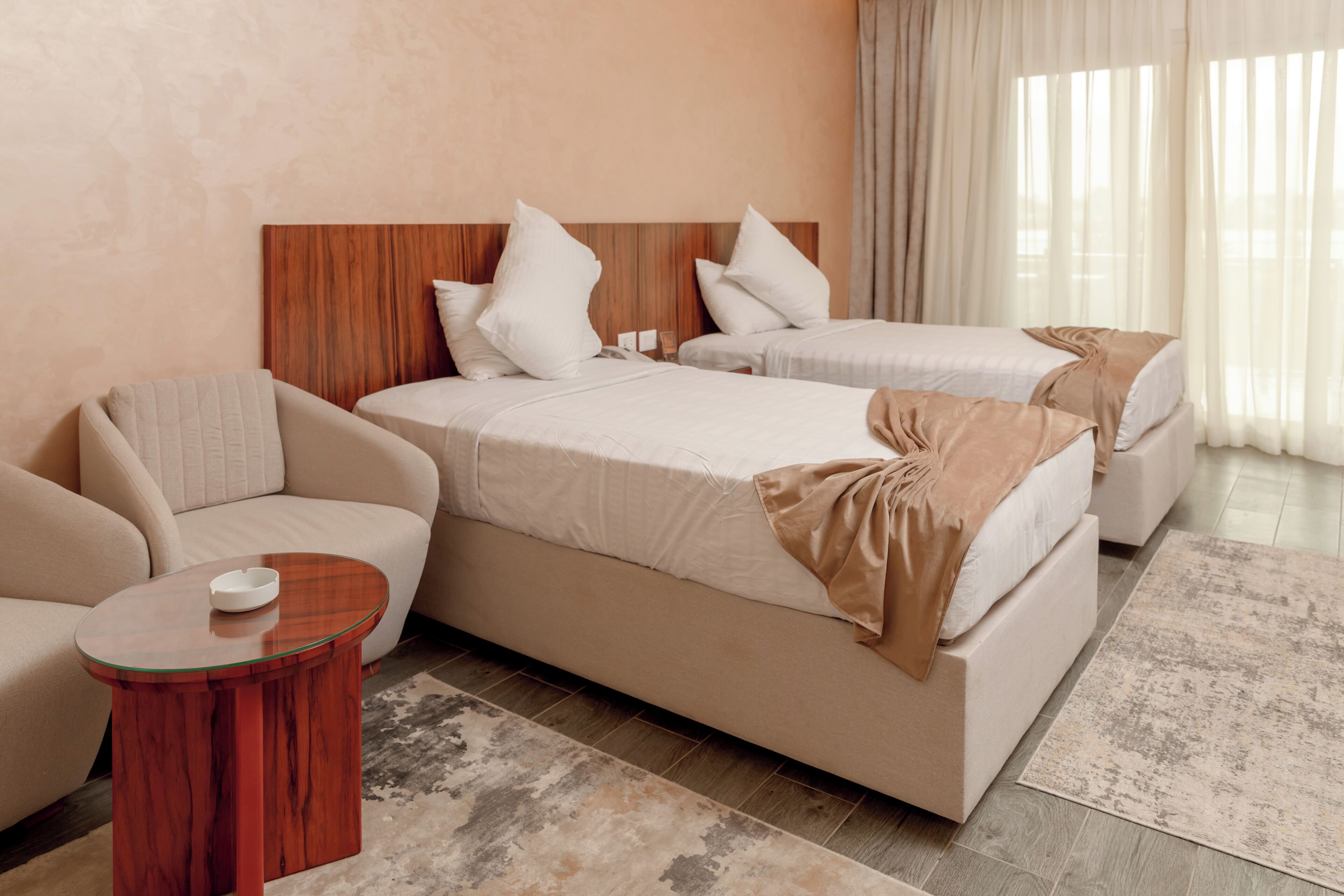 A hotel room with two beds, a sofa, a small table, and a large window with curtains.