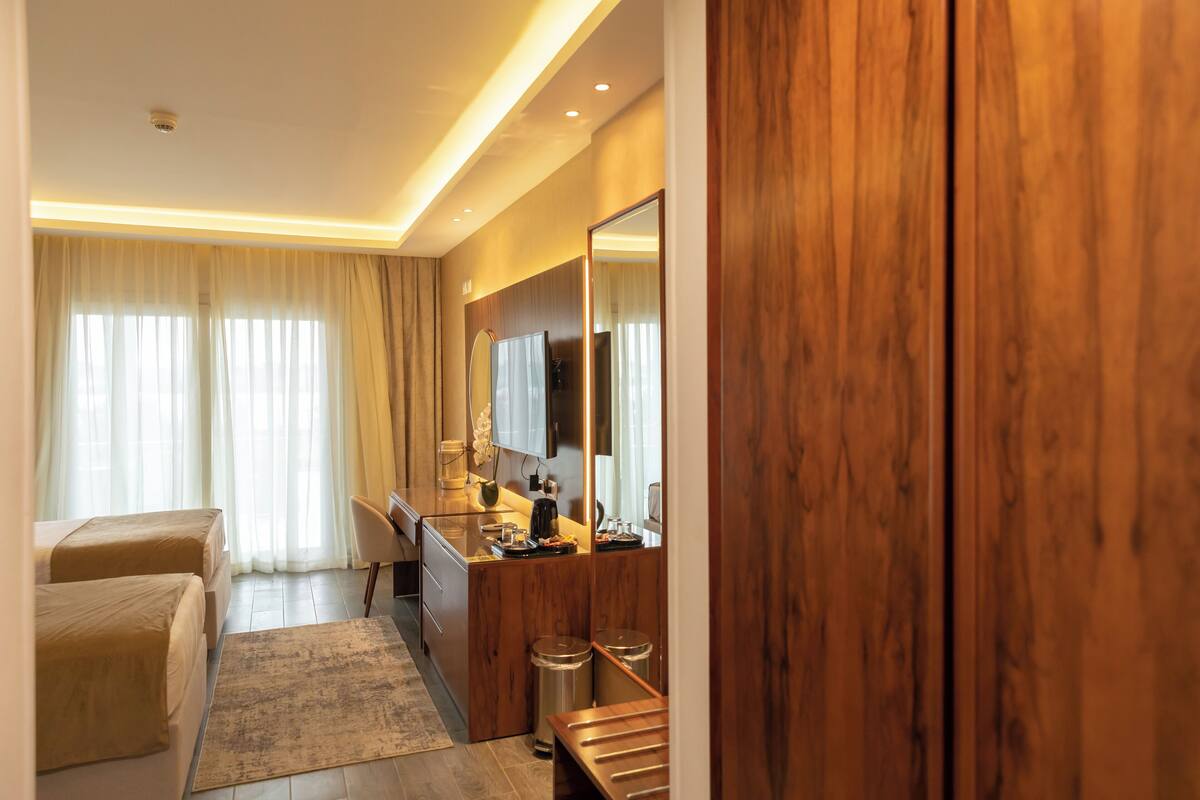 A hotel room with two beds, a wooden desk, a mirror, and a large window with curtains.