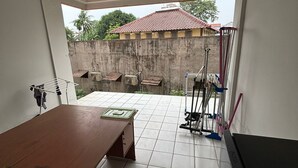 Una habitación con una gran mesa de madera, un armario negro y piso de baldosas. Se observa una pared de concreto y un portón metálico al fondo.