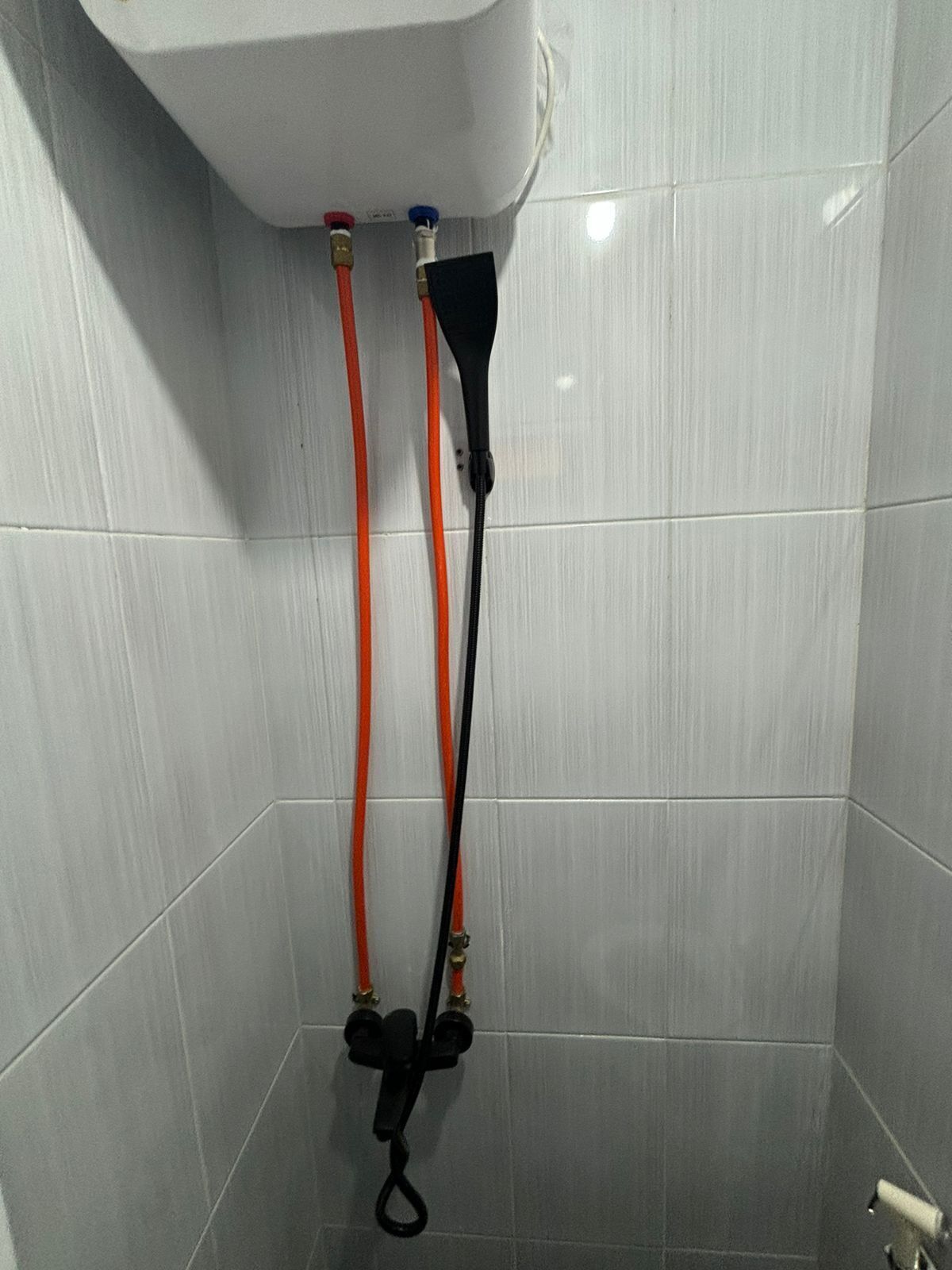 A tiled shower area with a white tank and two hoses, one red and one black, connected to it.