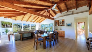 Une cuisine spacieuse avec des poutres en bois, un coin repas avec des chaises et une table, et de grandes fenêtres offrant une vue sur la verdure.