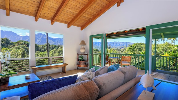 Un salon chaleureux avec un canapé, une table et une lampe. De grandes fenêtres offrent une vue sur les montagnes et la verdure.
