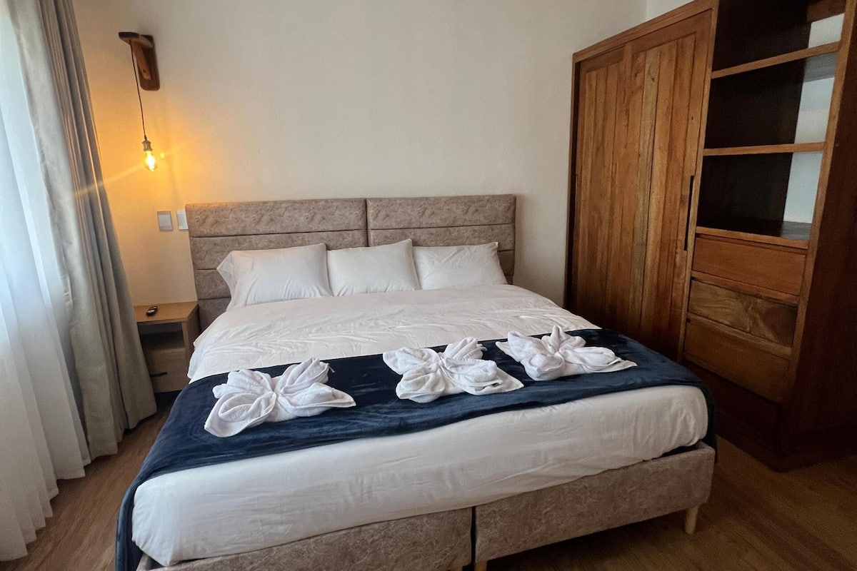 Una cama bien hecha con sábanas blancas y una manta azul, un armario de madera y una lámpara colgante.