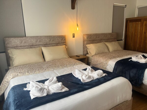 Habitación de hotel con dos camas, cada una con colcha y almohadas estampadas. Hay una mesita de noche con un control remoto, una lámpara de pared y un armario de madera.