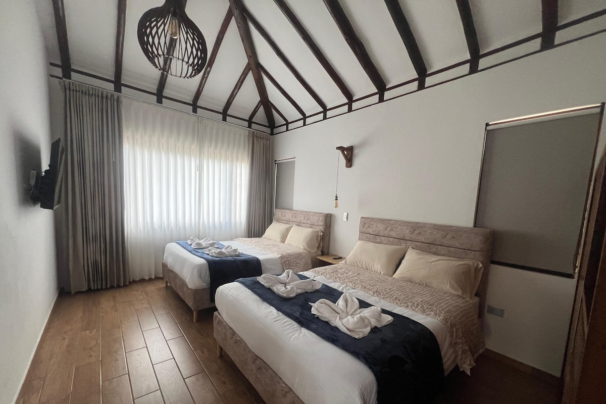 Habitación de hotel con dos camas, suelo de madera, un ventanal con cortinas, una lámpara de techo y un televisor de pantalla plana.