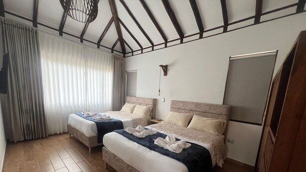 Ein Hotelzimmer mit zwei Betten, Holzboden, einem großen Fenster mit Vorhängen und einer Decke mit sichtbaren Balken.