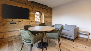 A modern living room with a wooden wall, a round table with chairs, a grey sofa, and a flat-screen TV mounted on the wall. - Lavish Apartment With Sauna (Lermoos)