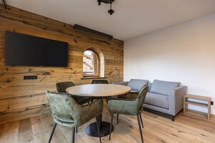 A modern living room with a wooden wall, a round table with chairs, a grey sofa, and a flat-screen TV mounted on the wall.