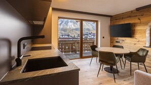 A modern kitchen with a sink, a dining area with chairs, a TV mounted on the wall, and a view of mountains through large windows. - Lavish Apartment With Sauna (Lermoos)