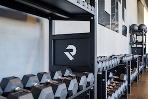 Un gimnasio con equipamiento de levantamiento de pesas, incluyendo mancuernas y discos, y un estante negro con un logotipo.