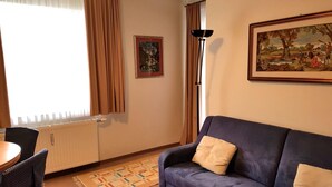 Una sala de estar con un sofá azul, una mesita, una silla, un radiador, una lámpara de pie y un cuadro en la pared.