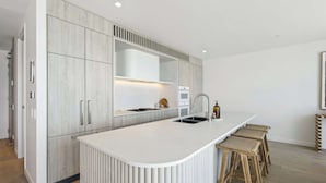 A modern kitchen with a white countertop, wooden cabinets, and a built-in stove. There are three wooden stools at the counter.