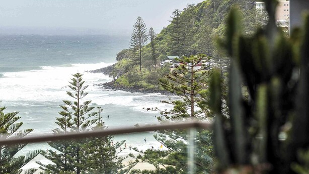 A coastal view with a balcony overlooking the ocean, a building with a glass facade, and a variety of trees.