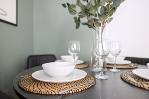A dining table set with white plates, bowls, and clear glassware, with woven placemats and a vase with green leaves. - Tranquil Pontcanna Mews (Cardiff)