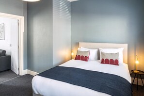 A neatly made bed with a white headboard, two pillows, and a dark blanket. There is a bedside table with a lamp, a door with a picture frame, and a grey sofa in the background. - Tranquil Pontcanna Mews (Cardiff)