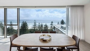 A dining area with a large table and chairs, overlooking a beach and ocean.