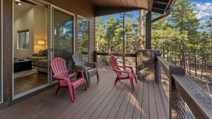 A wooden deck with Adirondack chairs, a glass door leading to a living area, and a view of trees.
