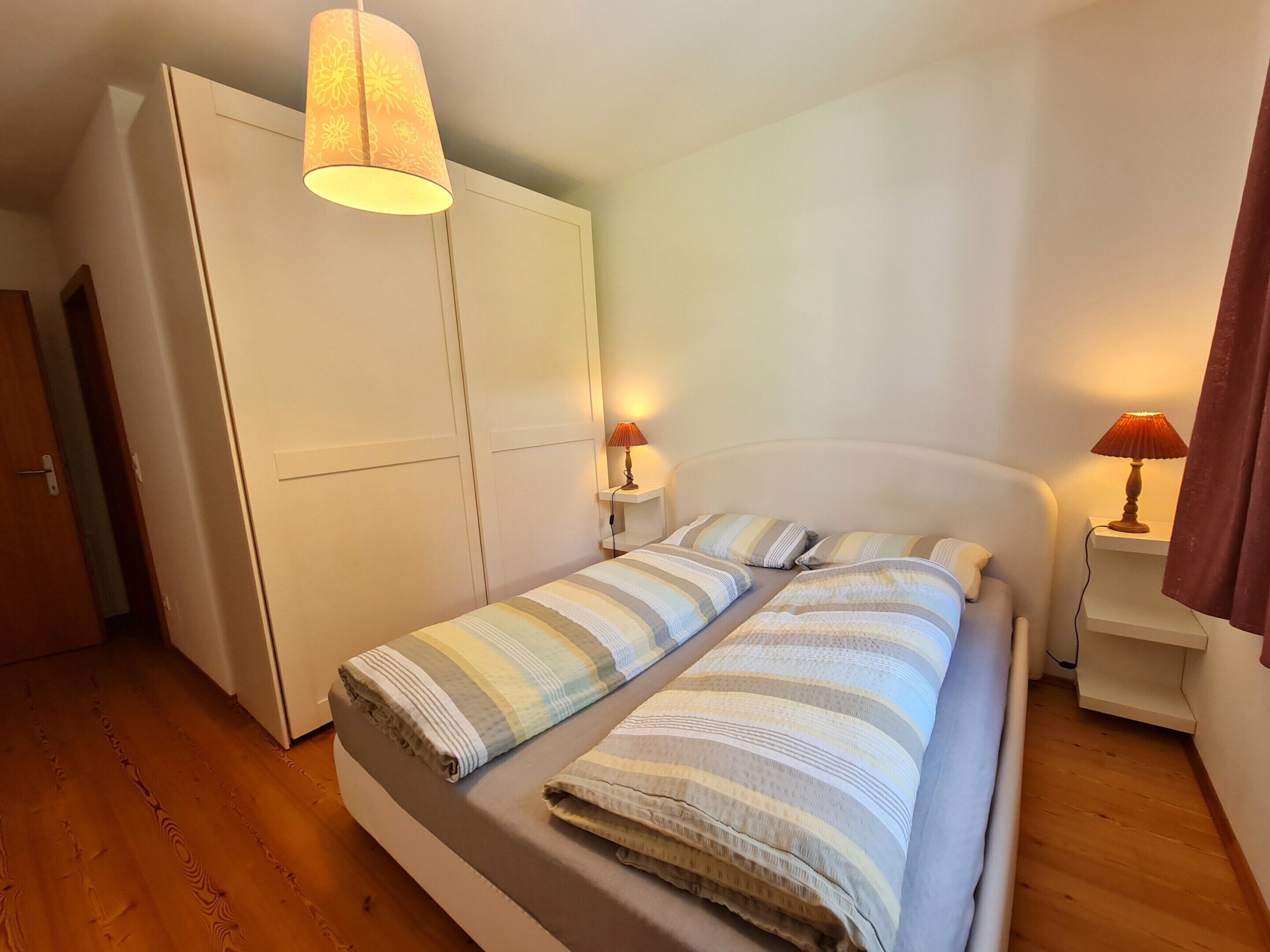 A single bed with striped bedding, two bedside lamps, a wardrobe, and a wooden floor.