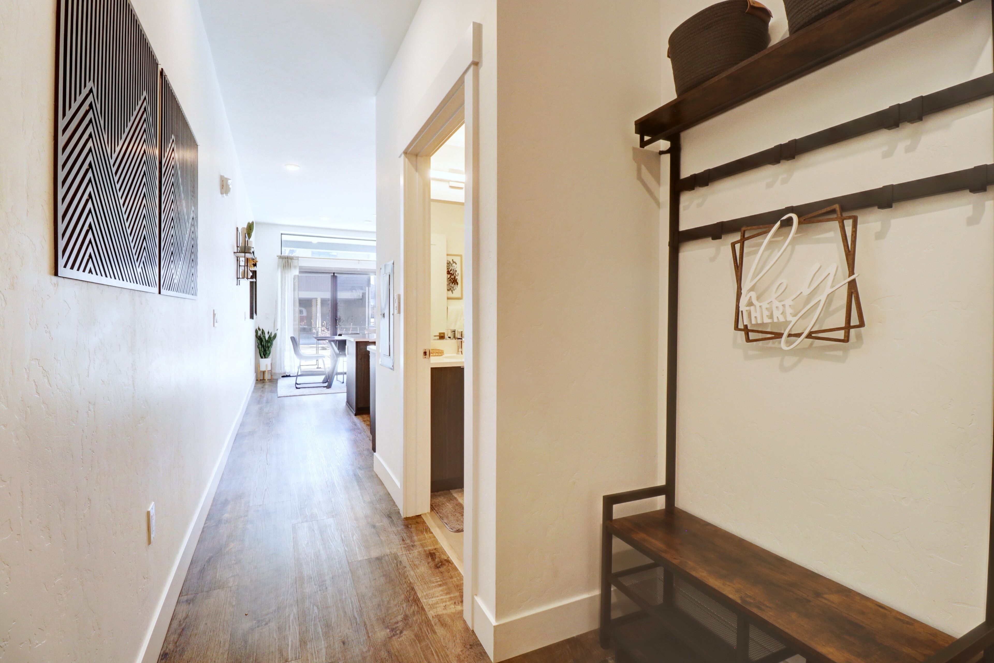 Un pasillo con suelo de madera, pared blanca con un panel decorativo y una estantería con cestas.