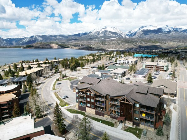 A modern hotel with multiple floors, a parking lot, and a view of a lake and mountains in the background.