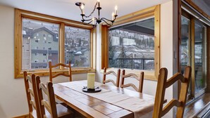 Comedor con mesa de madera, sillas y una araña. Amplias ventanas ofrecen vistas a edificios y un paisaje nevado.