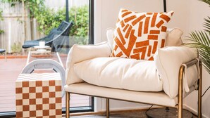 Ein modernes Wohnzimmer mit einer Couch, einem gemusterten Zierkissen, einem karierten Hocker und Blick auf eine Terrasses mit einem Stuhl und Pflanzen.