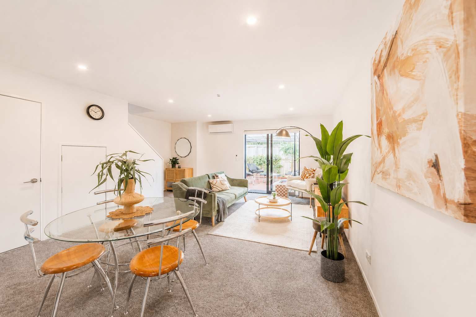 A modern living room with a glass dining table, a sofa, and a coffee table. There is a door leading to another room and a large window.