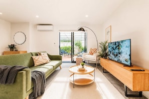 A modern living room with a green sofa, a coffee table, a TV on a wooden stand, and a view of the outdoors through a glass door.