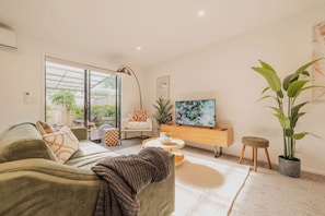 A modern living room with a sofa, coffee table, and TV. There is a large window, a plant, and a small stool.