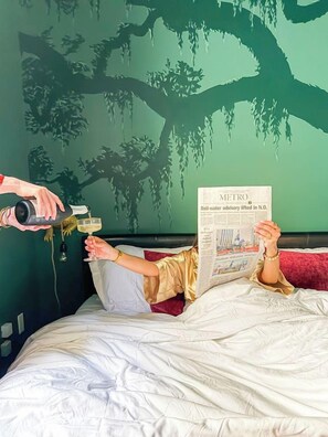 Eine Person liegt im Bett mit einer Zeitung, eine andere Person schenkt ein Getränk ein, und an der Wand ist ein Wandgemälde mit einem Baum.
