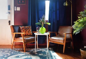A room with two leather chairs, a small table with a vase, a patterned rug, and a large window with blue curtains. - "La Lune Noire NOLA" (New Orleans)