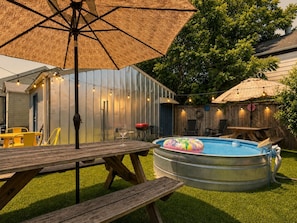 Un jardin avec une piscine, une table en bois, des chaises et un grand parasol.