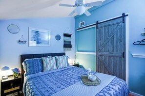 Un dormitorio con cama, ventilador de techo, repisa en la pared, puerta con acabado de madera y espejo adosado a la pared.