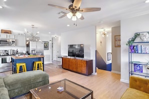Una cocina moderna con una isla central, un comedor con una mesa de centro de vidrio y una sala de estar con un sofá y una televisión.