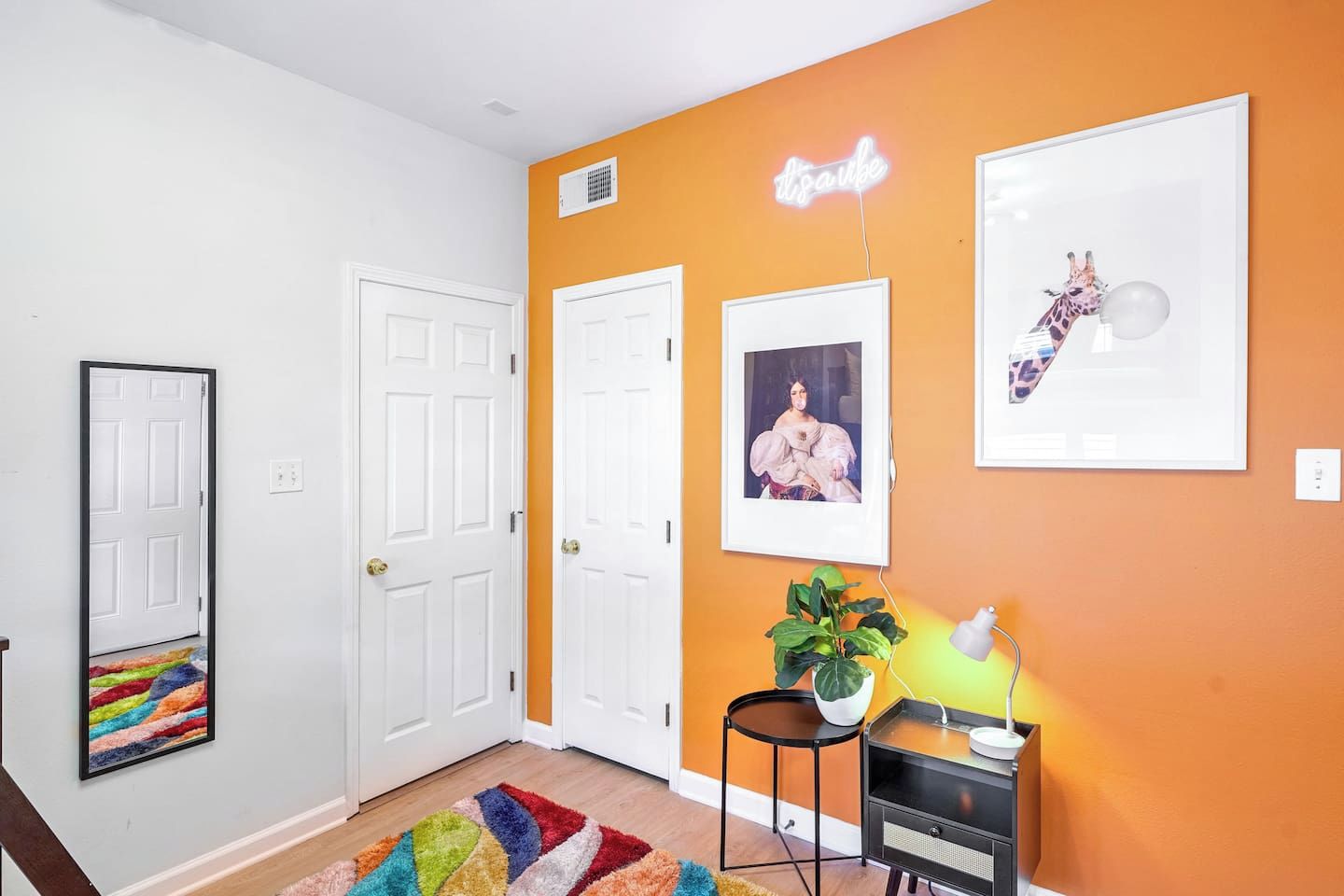 A hallway with white doors, a neon sign, framed artwork, a plant, and a small table with a lamp.