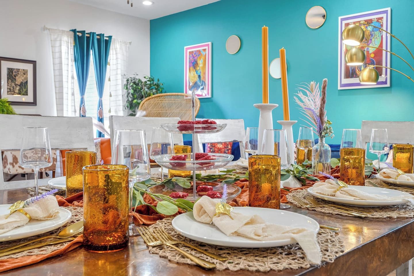 Mesa de jantar posta com copos, pratos e velas, com parede azul-turquesa e obras de arte ao fundo.