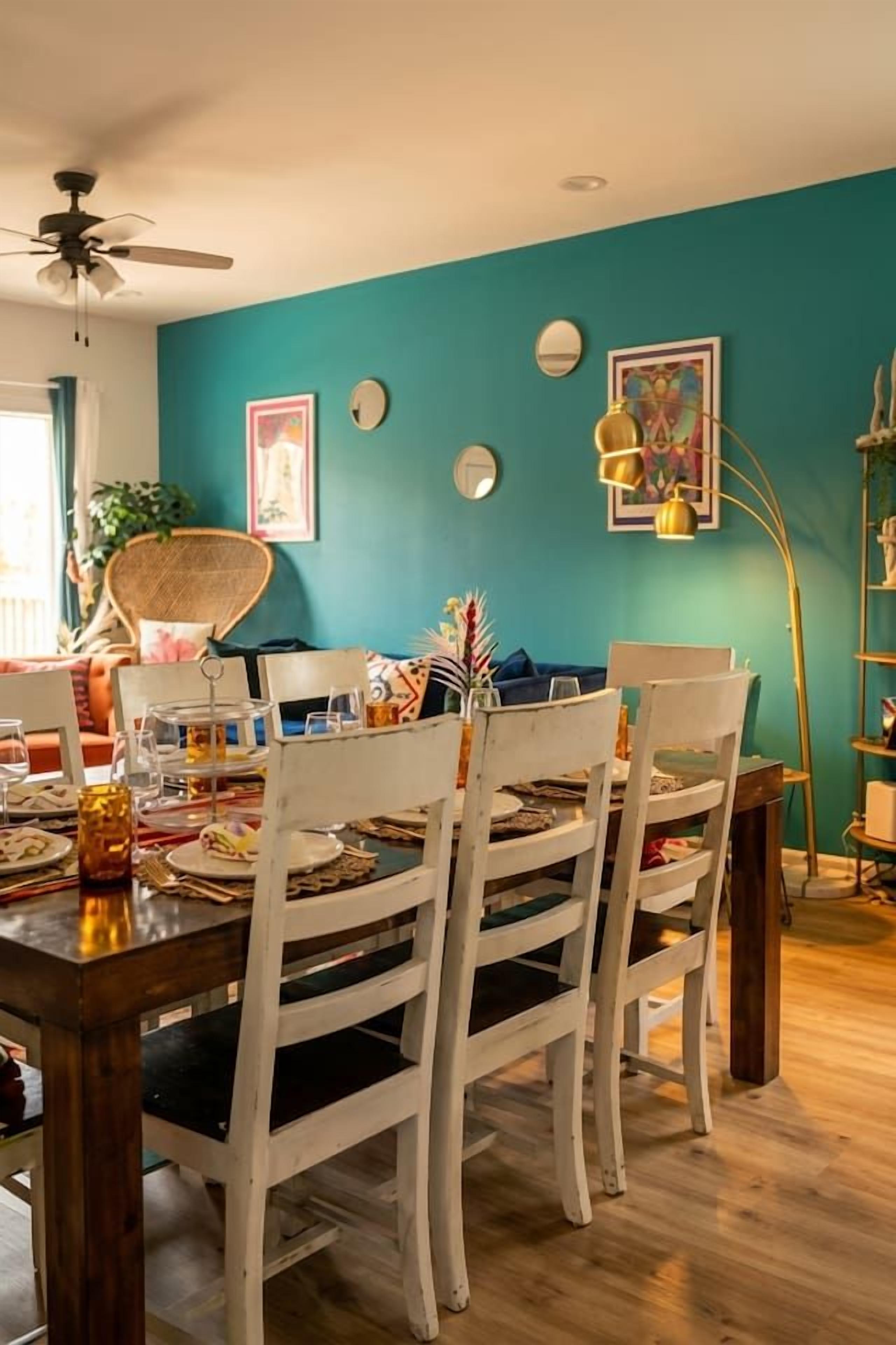 A dining area with a wooden table and white chairs, teal walls, and a ceiling fan.