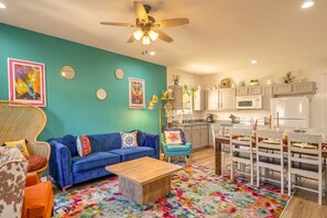 Ein Wohnzimmer mit einem blauen Sofa, einem Holztisch und einem farbenfrohen Teppich. Es gibt einen Essbereich mit einem weißen Tisch und Stühlen, sowie eine Küche mit weißen Schränken und Geräten.