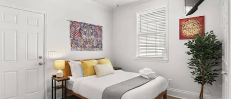 A bedroom with a bed, bedside table, lamp, artwork, window, and a potted plant.