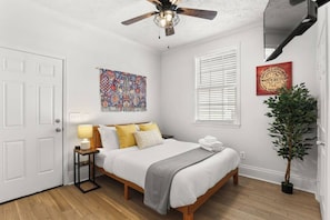 A bedroom with a bed, bedside table, lamp, artwork, window, and a potted plant. - Adorable 3BR, Cowboy Pool & Porch in Mid-City (New Orleans)