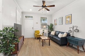 A living room with a ceiling fan, a sofa, a yellow armchair, a coffee table, and a TV. - Adorable 3BR, Cowboy Pool & Porch in Mid-City (New Orleans)