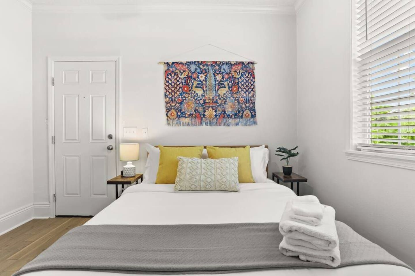 Een slaapkamer met een bed, een deur, een wanddecoratie, een nachtkastje met een lamp en een raam met jaloezieën.