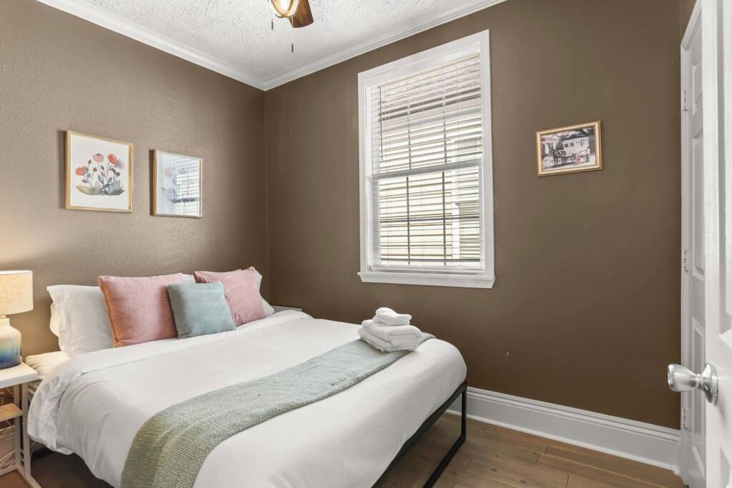 A bedroom with a bed, pillows, a bedside table, a lamp, a window with blinds, and wall art.