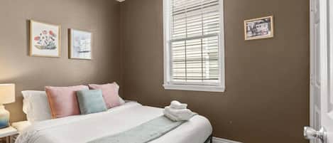 A bedroom with a bed, pillows, a bedside table, a lamp, a window with blinds, and wall art.