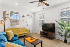 A living room with a yellow sofa, a blue sofa, a wooden coffee table, a TV stand, and a ceiling fan.