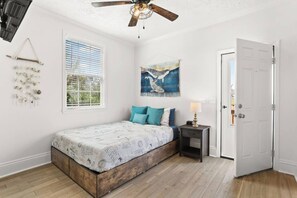 A bedroom with a bed, a nightstand, a ceiling fan, a window with blinds, a door, and wall art.